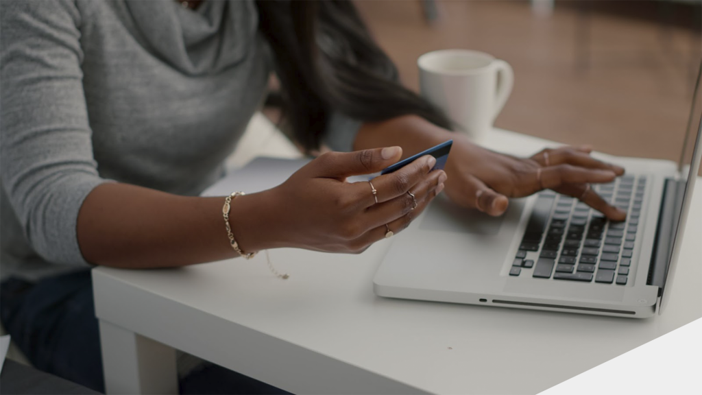 Mulher negra sentada à uma mesa com um notebook segurando um cartão e digitando com a outra mão, fazendo compras online.