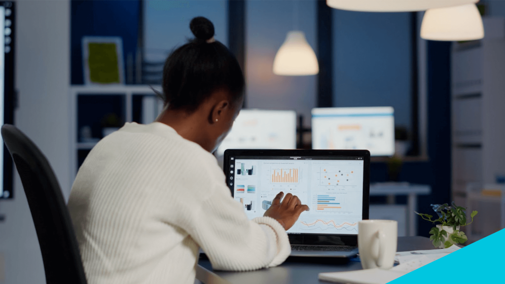 Mulher negra sentada à mesa de trabalho, analisando gráficos de dados coletados no computador.