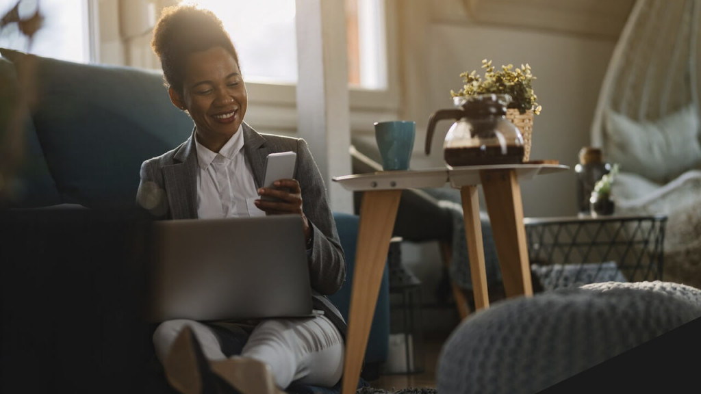 Mulher negra sorrindo, com um notebook no colo e um celular na mão, ao lado de uma mesa com café.