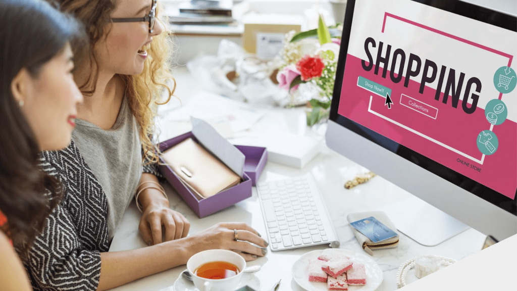 Duas mulheres, sentadas à uma mesa com chás e doces, olhando para a página de um e-commerce no computador. Na página, está escrito “Shopping”.