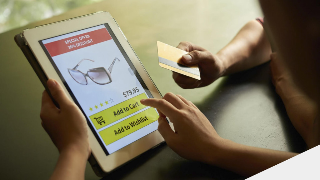 Mão segurando um tablet exibindo página de compra de um óculos, com outra mão esticada com um cartão de crédito ao lado.